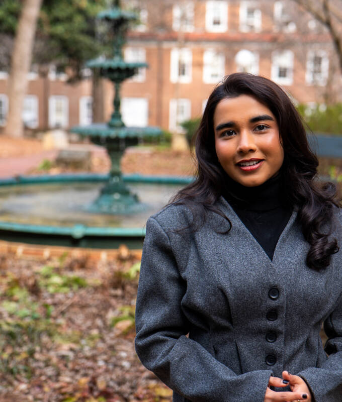 Female student in front of a historical fountain with foilage around.